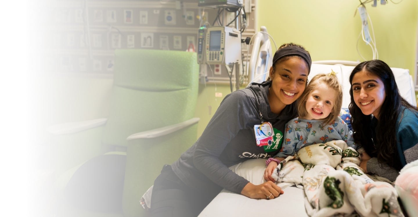 two nurses smile on either side of a pediatric patient smiling in her hospital bed