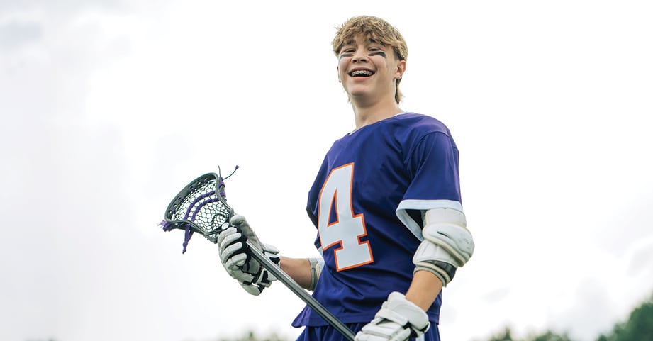 Teen lacrosse player smiles with lacrosse stick and gear.
