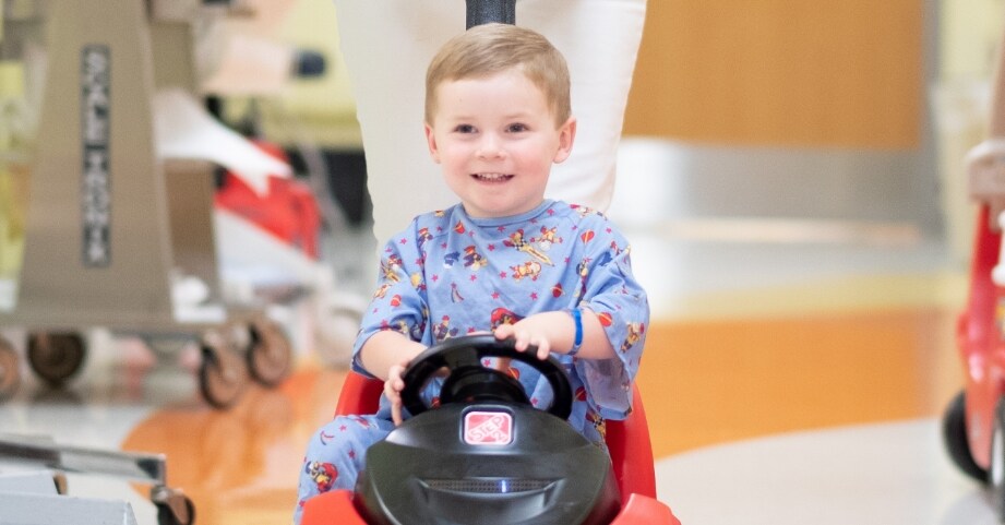 A happy boy wheels through hospital in toy car.