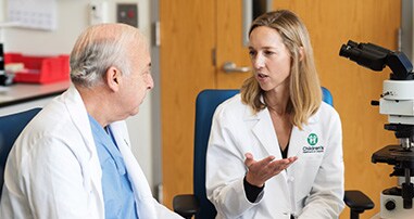 A male and a female doctor, each in a white coat, are sitting in a lab talking