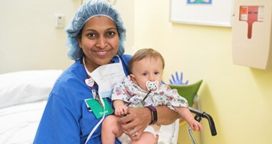 A pediatric heart surgeon wearing scrubs smiles while holding a boy patient before his surgery
