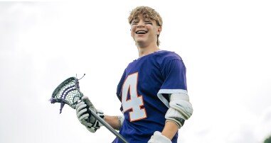 Teen lacrosse player smiles with lacrosse stick and gear. 