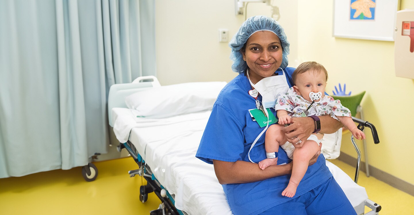 A pediatric heart surgeon wearing scrubs smiles while holding a boy patient before his surgery