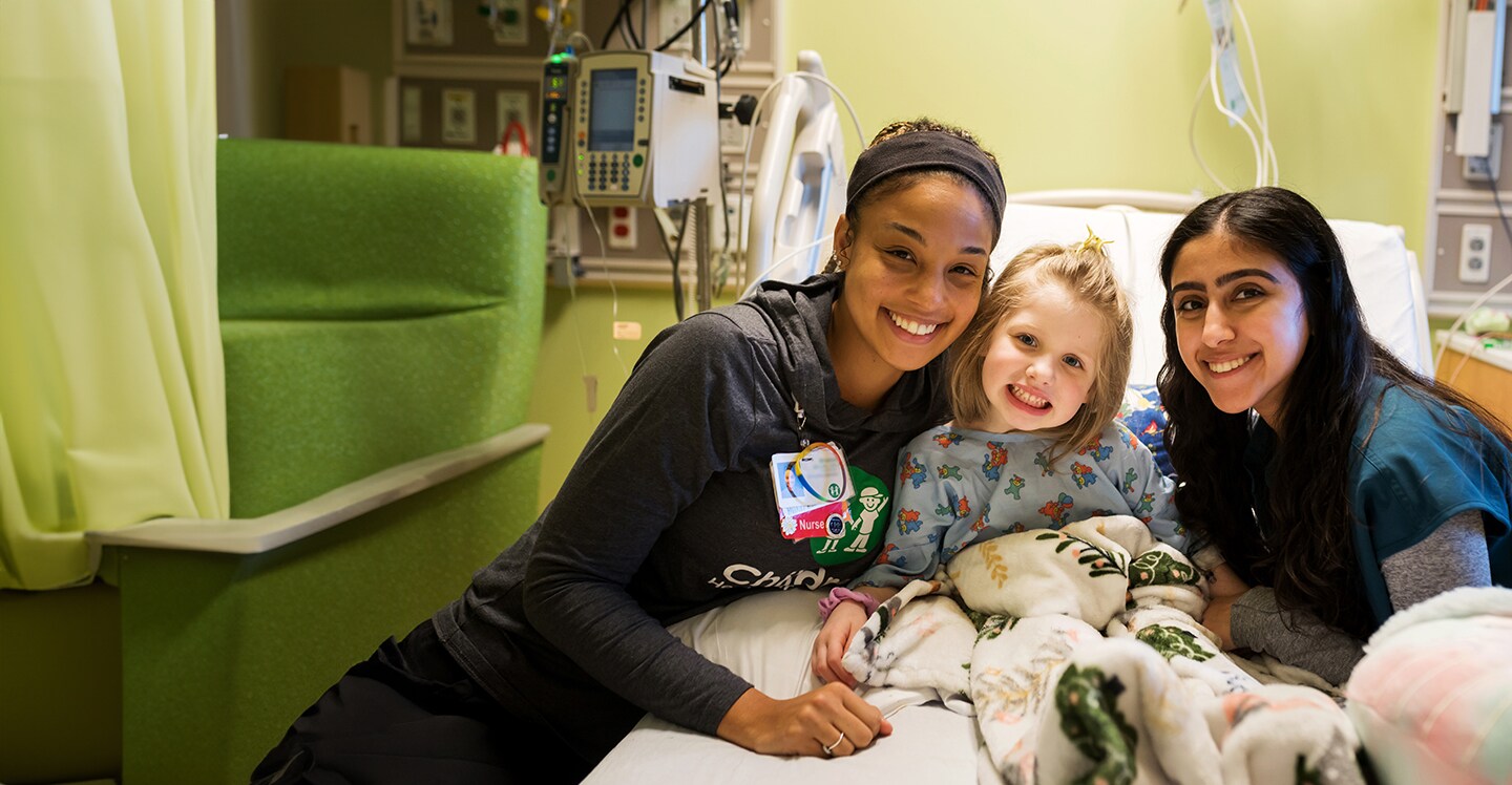 Young girl patient smiling in her hospital bed with two female nurses smiling on either side of her.