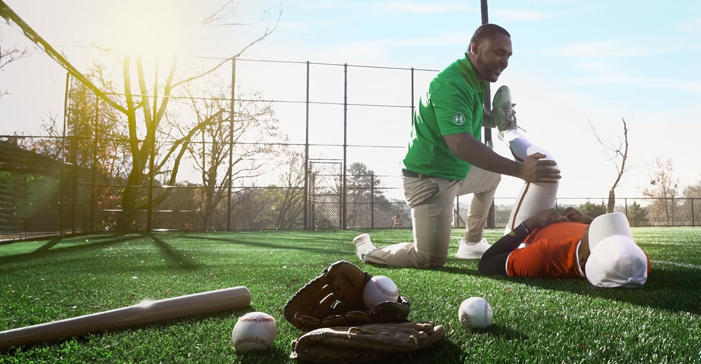 Children's athletic trainer on field helping teen baseball player stretch before a game.