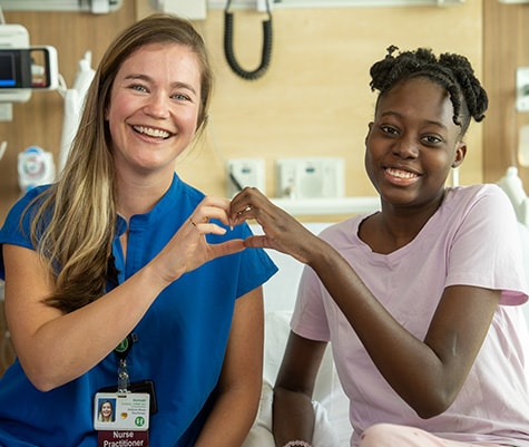 A female nurse sits with a patient on her bed, putting their hands together to form a heart while smiling.