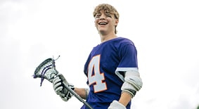 Teen lacrosse player smiles with lacrosse stick and gear. 